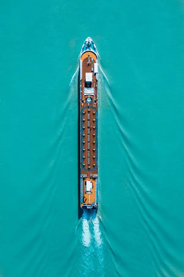 Aerial view of a large ship moving through turquoise water, with visible wake patterns and deck structures, representing maritime travel and an oceanic voyage.