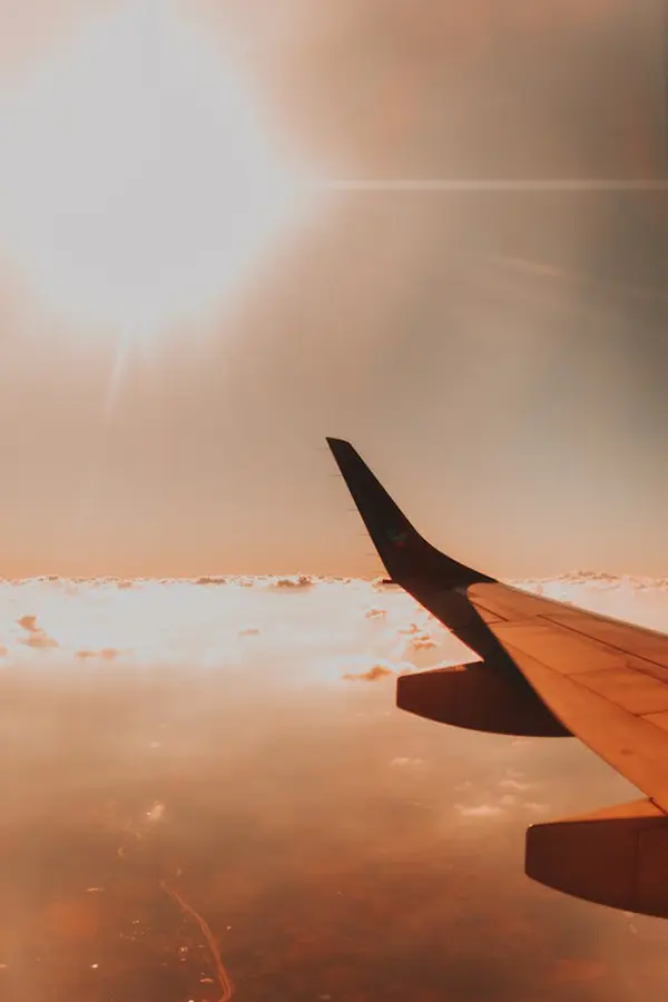 Airplane wing photographed during flight, framed by a gradient sky with warm orange hues, illuminated clouds, and a glowing sunrise, capturing the peaceful essence of aerial travel.