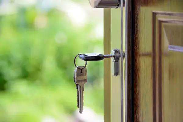 Close-up of a wooden door with a key inserted in the lock, slightly open, revealing a blurred green outdoor background, highlighting home security and entry.
