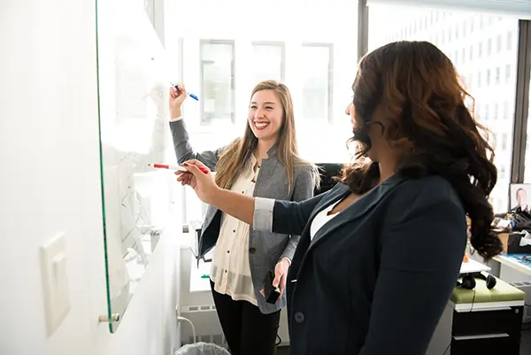 Two women in a modern office collaboratively using a glass board for a brainstorming session, with natural light and office furniture in the background.