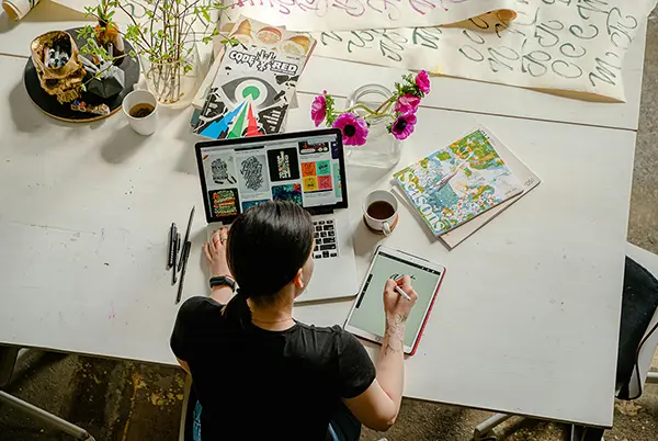 Creative workspace with a person using a tablet and stylus for graphic design, a laptop displaying designs, and artistic elements like flowers, a magazine, and writing tools.
