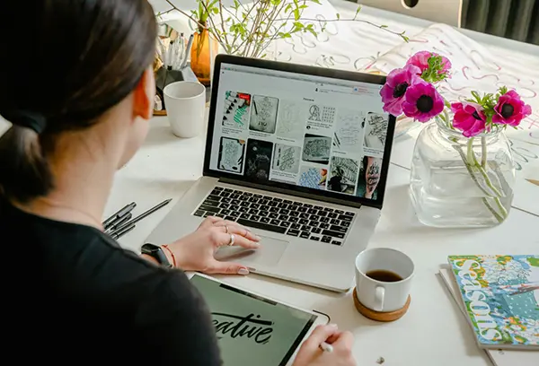 Person at a table in a creative workspace, using a laptop with typography designs and a tablet displaying the word creative, surrounded by a coffee cup, a magazine titled Seasons, and pink flowers in a vase.