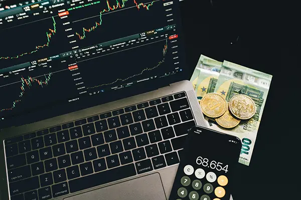 Financial trading setup with a laptop screen showing Ethereum cryptocurrency charts, a smartphone displaying a calculator app reading 68.654, and Bitcoin coins alongside Euro banknotes illustrating digital and traditional finance integration.