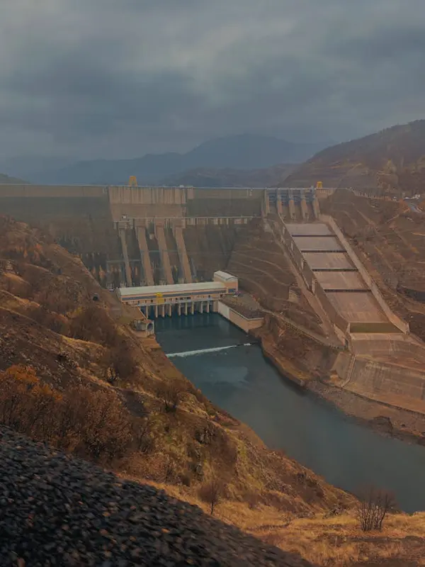 Large dam with vertical spillways releasing water into a river set in a mountainous area with sparse vegetation and an overcast sky, emphasizing hydroelectric power and water management systems.
