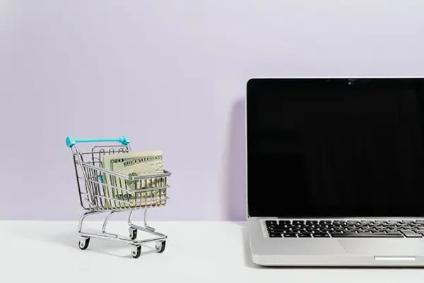 Small shopping cart filled with US dollar bills beside an open laptop on a white surface with a pastel purple background, highlighting the concept of e-commerce and digital transactions.