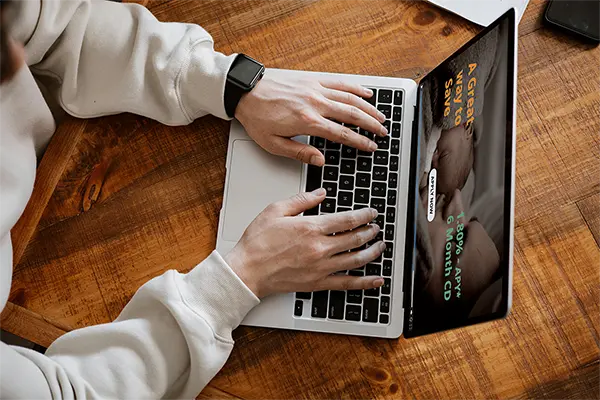 Person typing on a laptop displaying a financial advertisement for a 6-month CD with a 1.80 percent APY and an Apply Now button on the screen, promoting savings opportunities.