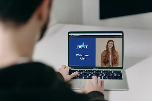 Laptop screen showing a personalized welcome message from First Credit Union, with a blue background, text reading Welcome John, and an image of a smiling person highlighting customer onboarding in online banking.