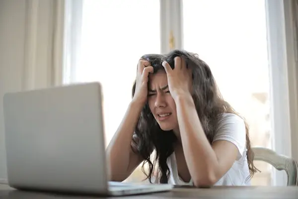 Person with long dark hair sitting at a table in front of a laptop, visibly stressed with their hands on their head, in a bright home office setting with a window in the background.