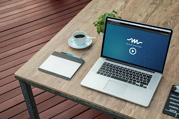 Workspace setup with a laptop displaying the mediawide logo, a graphics tablet, a coffee cup, a plant, and a clapperboard, ideal for media production and digital content creation