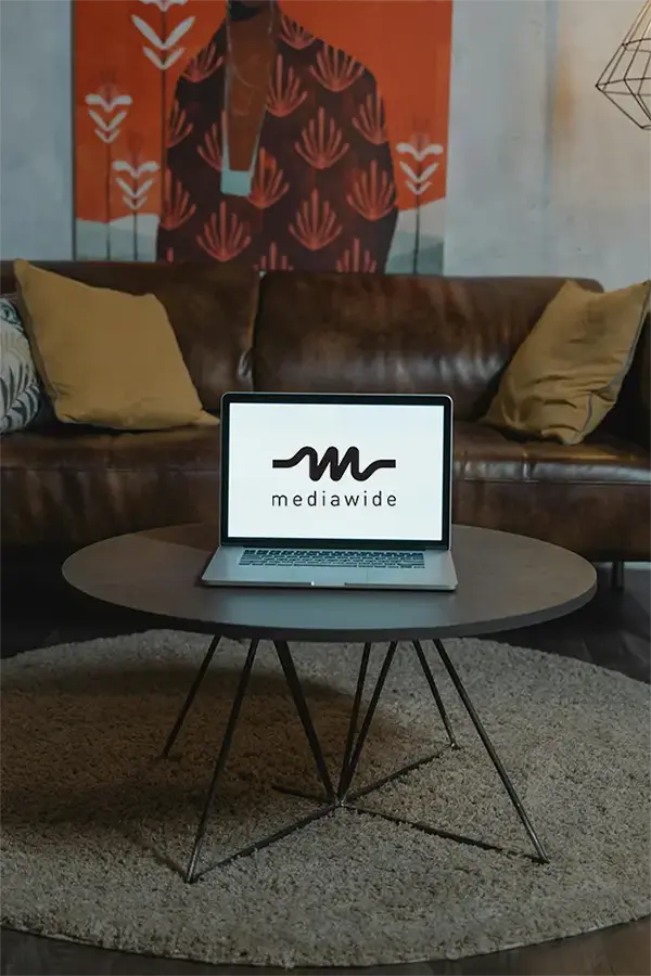 Laptop with mediawide displayed on the screen, placed on a round wooden table with a textured rug in a cozy workspace featuring a brown leather couch, cushions, and abstract wall art.