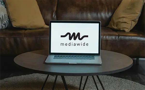 Laptop on a round coffee table in a living room, featuring the mediawide logo with a stylized wave design on the screen, emphasizing a blend of comfort and professional aesthetics.