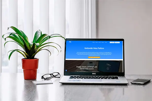 Workspace with a DELL laptop showing the Mediawide Video Platform website. Features include a blue and grey color scheme, text promoting personalized video marketing, and a call-to-action button, with additional items like a potted plant and smartphone.