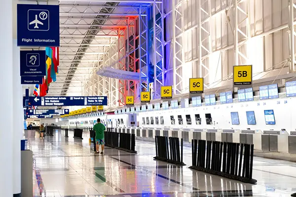 Modern airport terminal with check-in counters, international signage for customs and arrivals, hanging international flags, and vibrant blue and red lighting. Two individuals are visible navigating the spacious area.