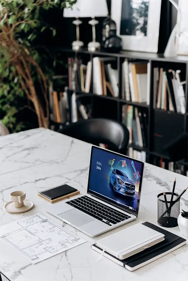 Modern workspace with a laptop on a marble table displaying a car model and Hello, John greeting, surrounded by a cup, notebooks, a pen holder, and a blueprint, with books and decorations in the background.