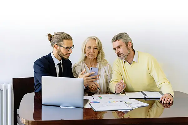 Three individuals in a business meeting seated at a table, focused on a smartphone. The table displays a laptop, colorful charts, and handwritten notes, reflecting collaboration and data analysis.