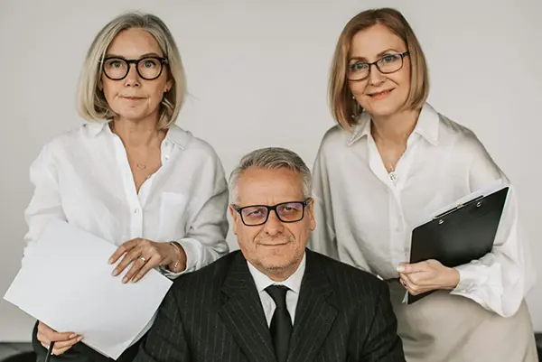 Professional team of three featuring a man in a dark pinstripe suit and two women in white blouses, holding documents, symbolizing business collaboration and teamwork.