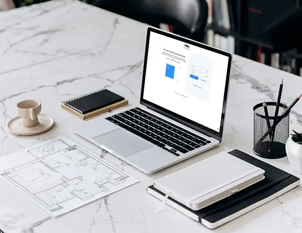 Workspace setup with a laptop open to a project exploration webpage on a marble table, surrounded by a coffee cup, notebooks, an architectural floor plan, and a pencil holder, with bookshelves and a plant in the background.
