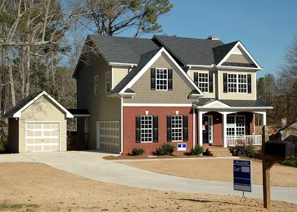 Two-story suburban house with red brick and gray siding, a detached garage, curved driveway, and an available lot sign, surrounded by a winter setting with dry grass and leafless trees.