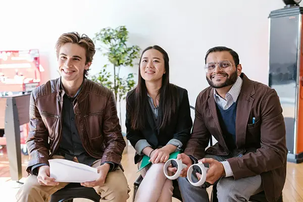 Three individuals seated in a well-lit indoor setting, holding items like a green folder, papers, and white circular devices, suggesting a casual professional collaboration in a tech-focused context.
