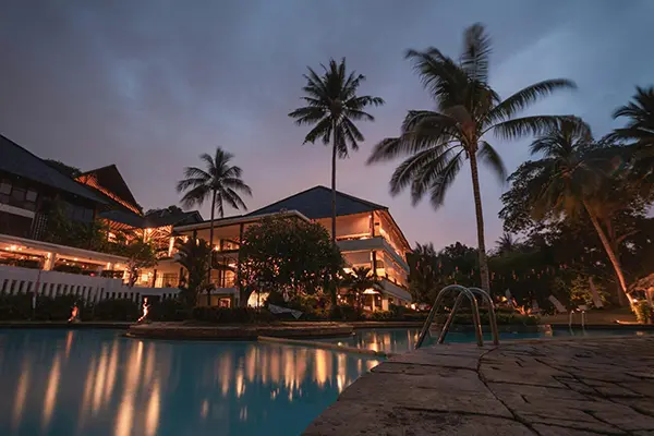View of a tropical resort at dusk with a warmly lit multi-story building, tall palm trees, and a calm swimming pool reflecting the evening sky.