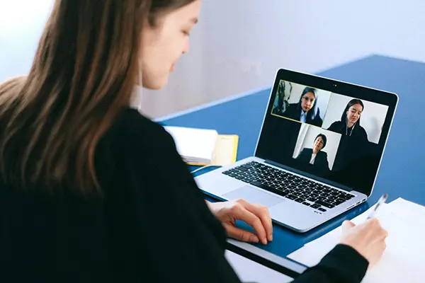 Woman participating in a video call on a laptop with three participants displayed in individual windows, seated at a desk with a notepad, papers, and a pen, symbolizing remote work and online collaboration.
