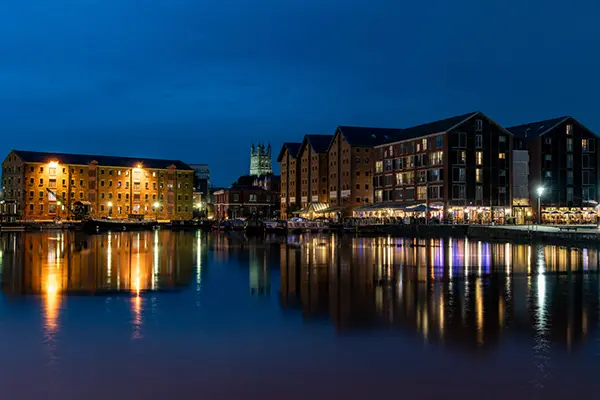 Nighttime waterfront scene with historical brick buildings, warm glowing lights, and a cathedral-like structure in the background, reflecting on calm water.